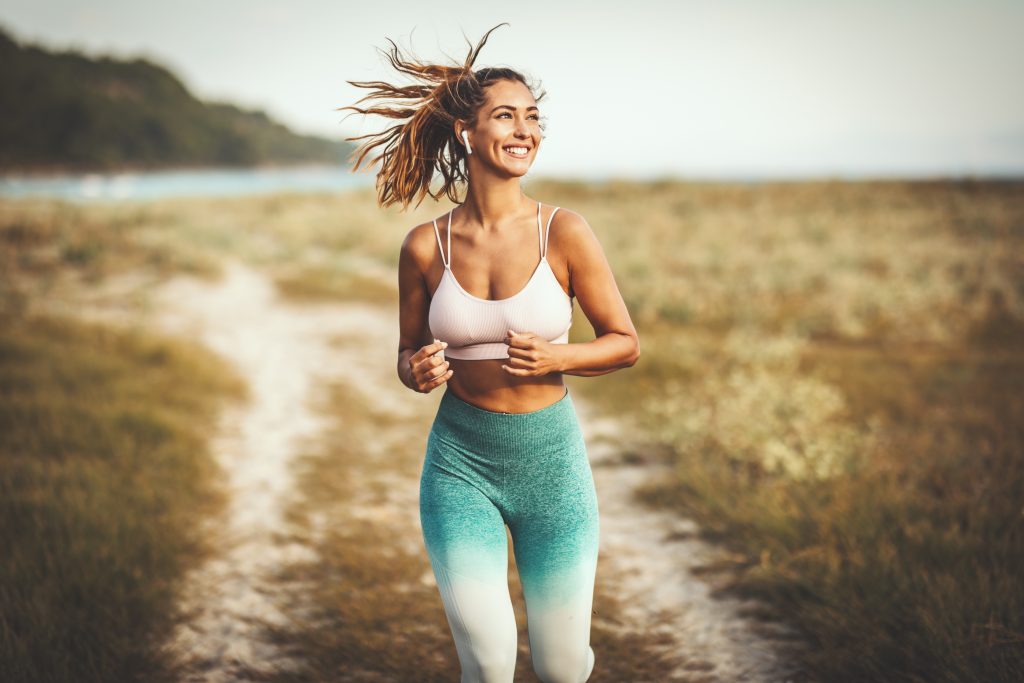 A beautiful smiling happy woman is running along the path near the sea and enjoying in summer sunny day.
